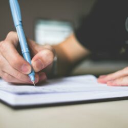 person holding blue ballpoint pen writing in notebook